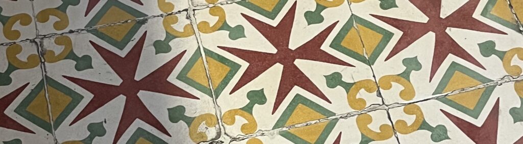 A photo showing detail of a tiled floor with a pattern with the wine red cross of Malta in the middle and decorative and geometric pattern in muted green and yellows with off white background. Photo taken in Valletta, Malta.